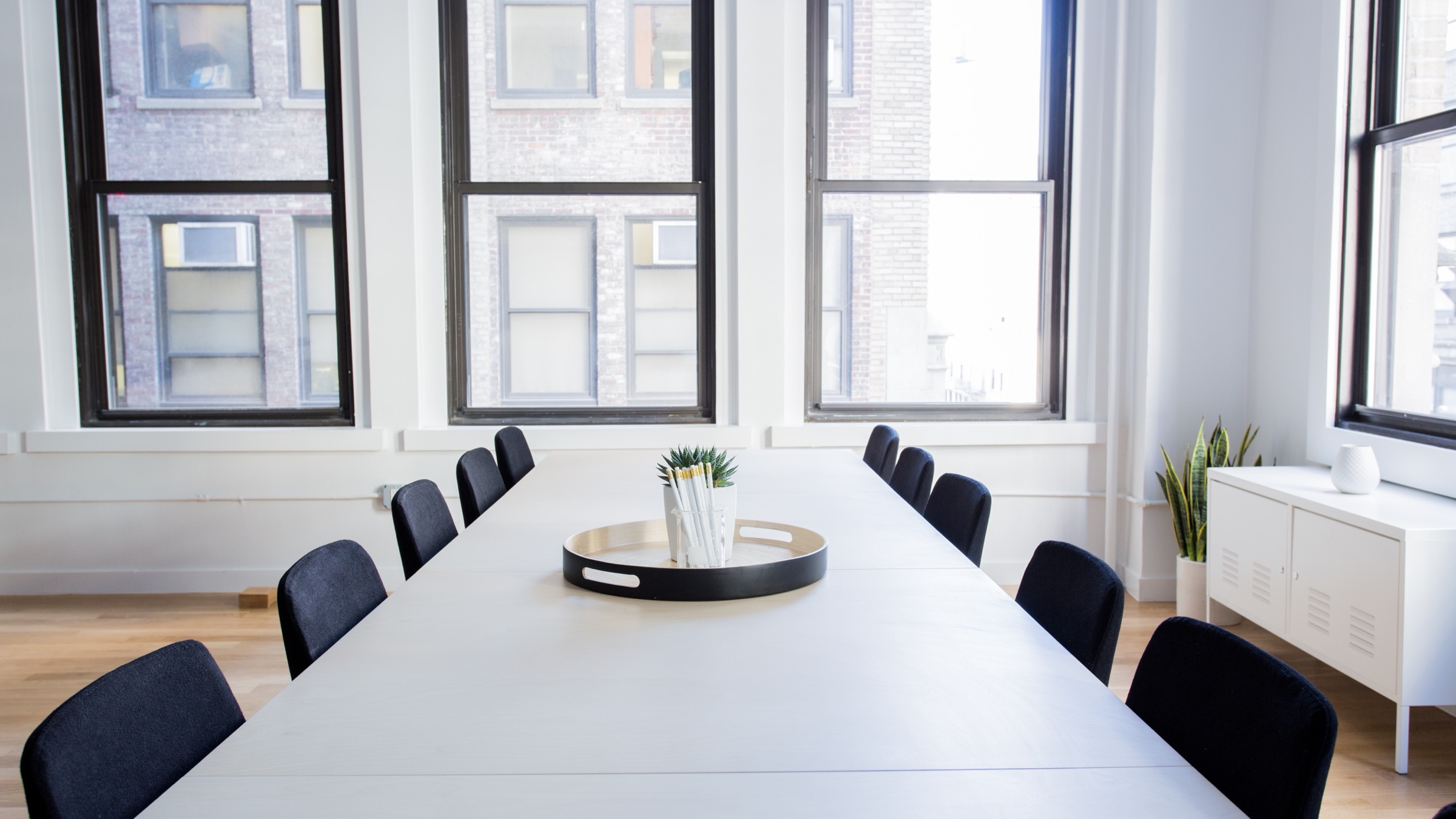 An empty white meeting room with a long table and twelve chairs, lit by tall windows.