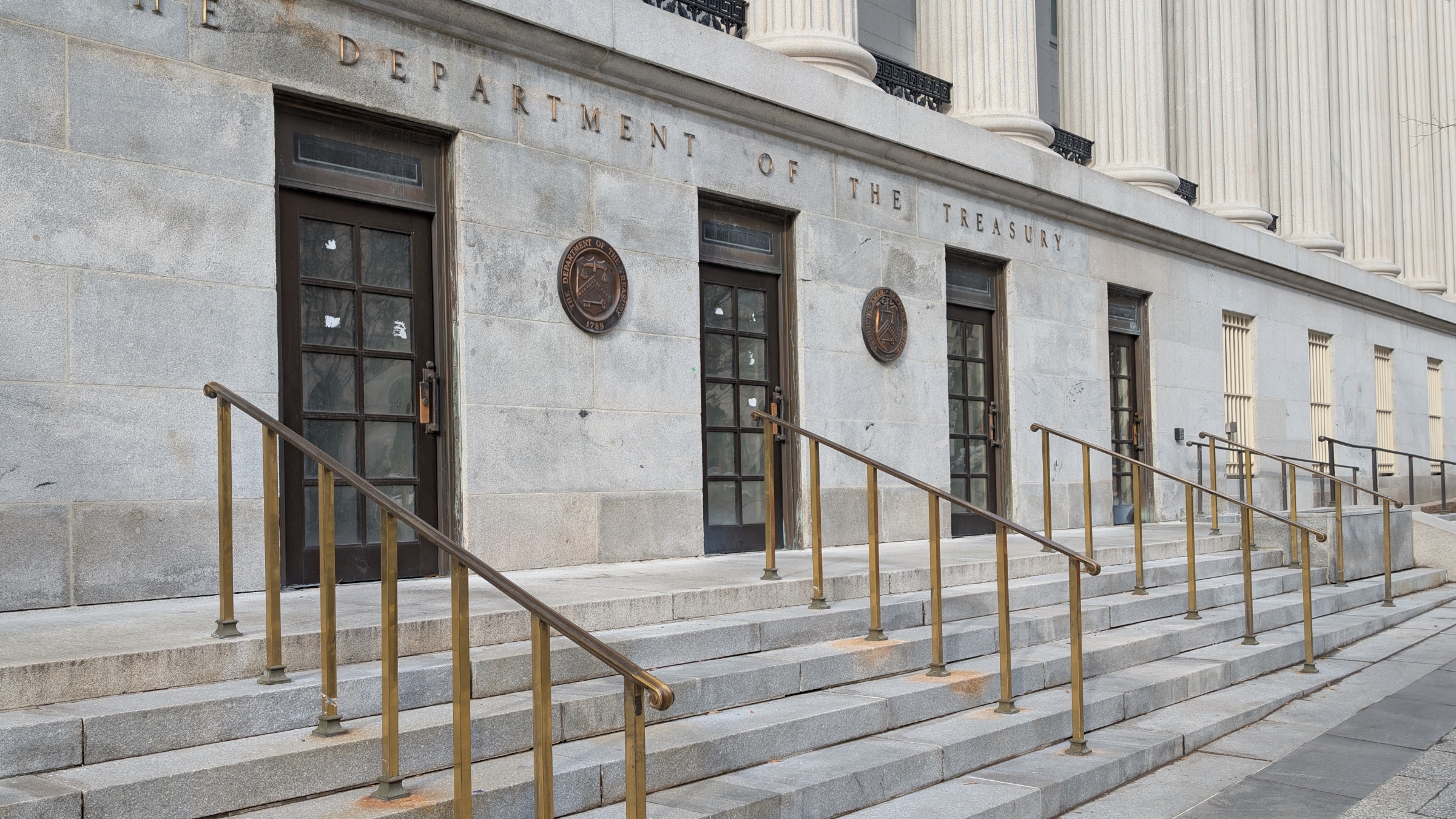 The side entrance of the United States Department of the Treasury building in Washington, with the department's name carved in stone above the doors and two bronze seals on the facade.