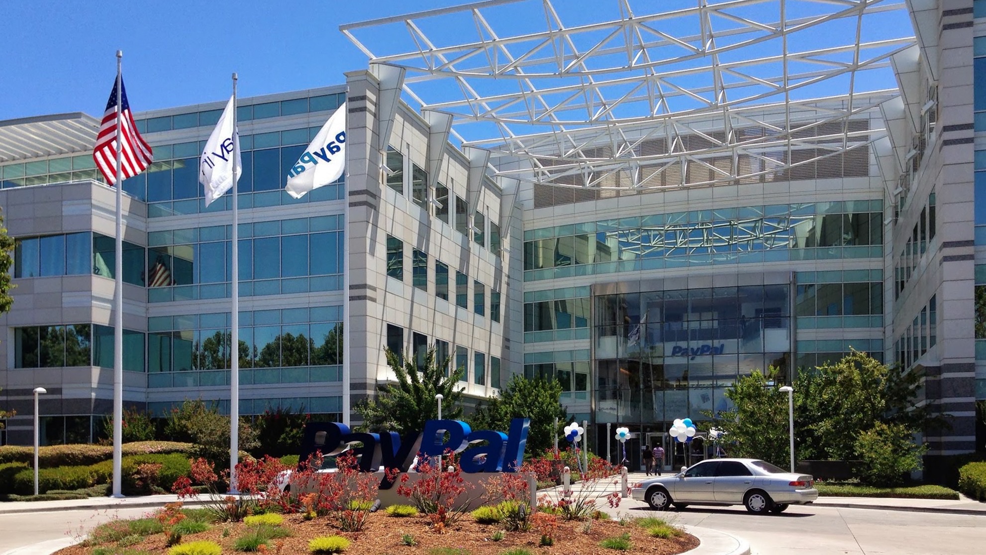 PayPal's San Jose headquarters, a glass office building with the company's logo on a ground-level sign and flags at the entrance.