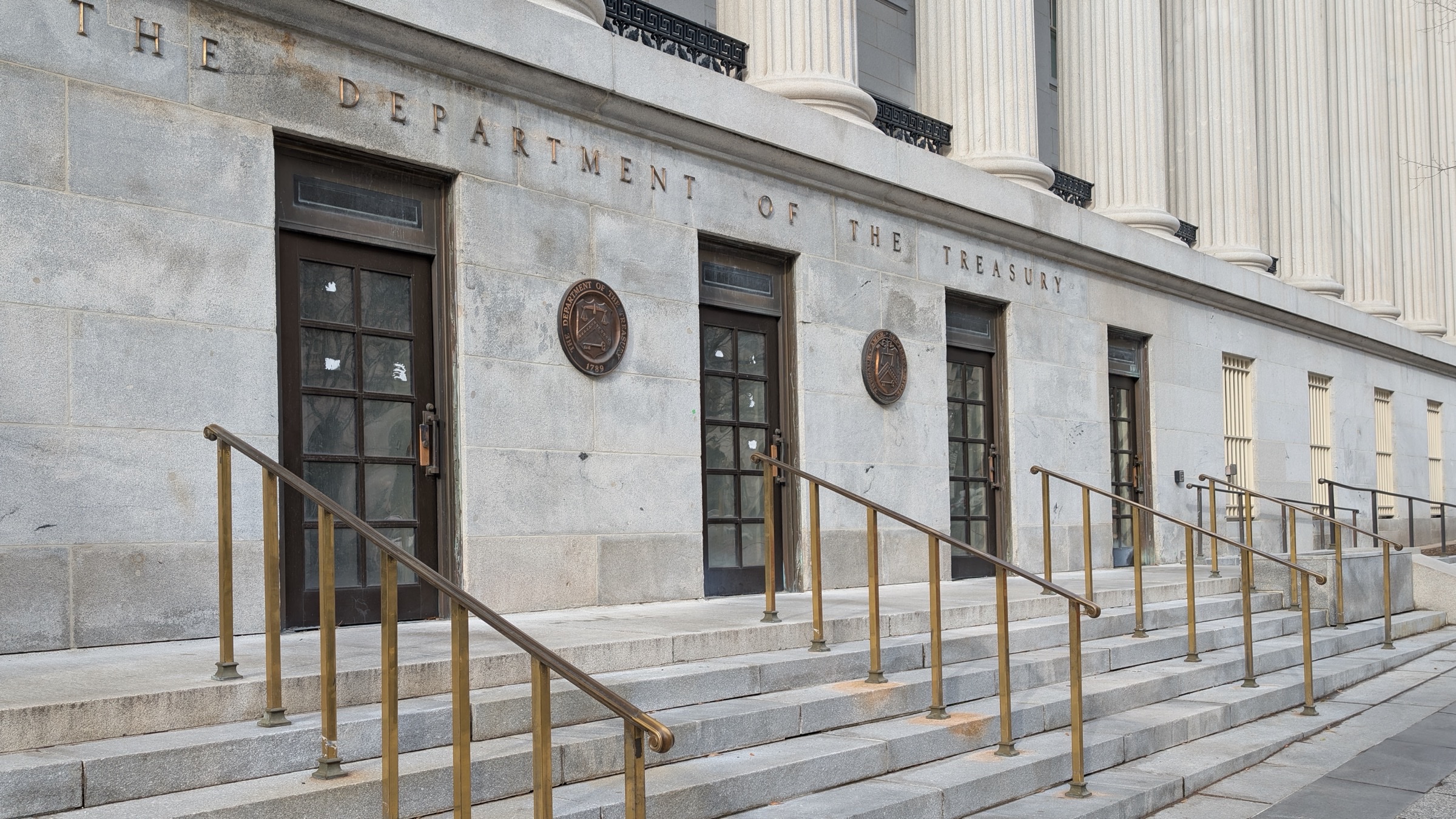 The engraved stone facade of the US Treasury building in Washington reading "The Department of the Treasury", with brass handrails on the entrance steps.