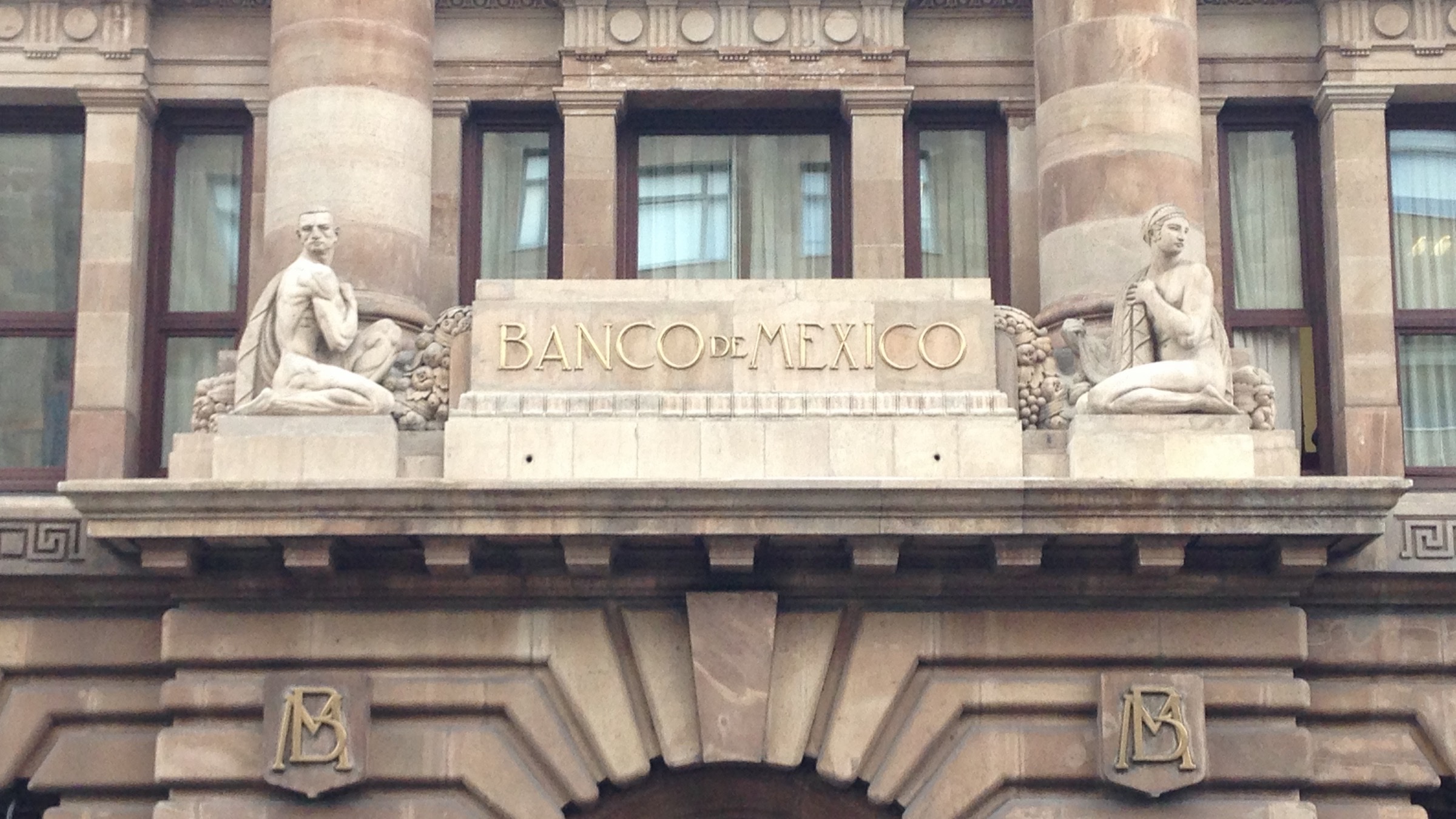 The stone facade of the Bank of Mexico in Mexico City, with "Banco de México" engraved above the entrance between two carved figures.