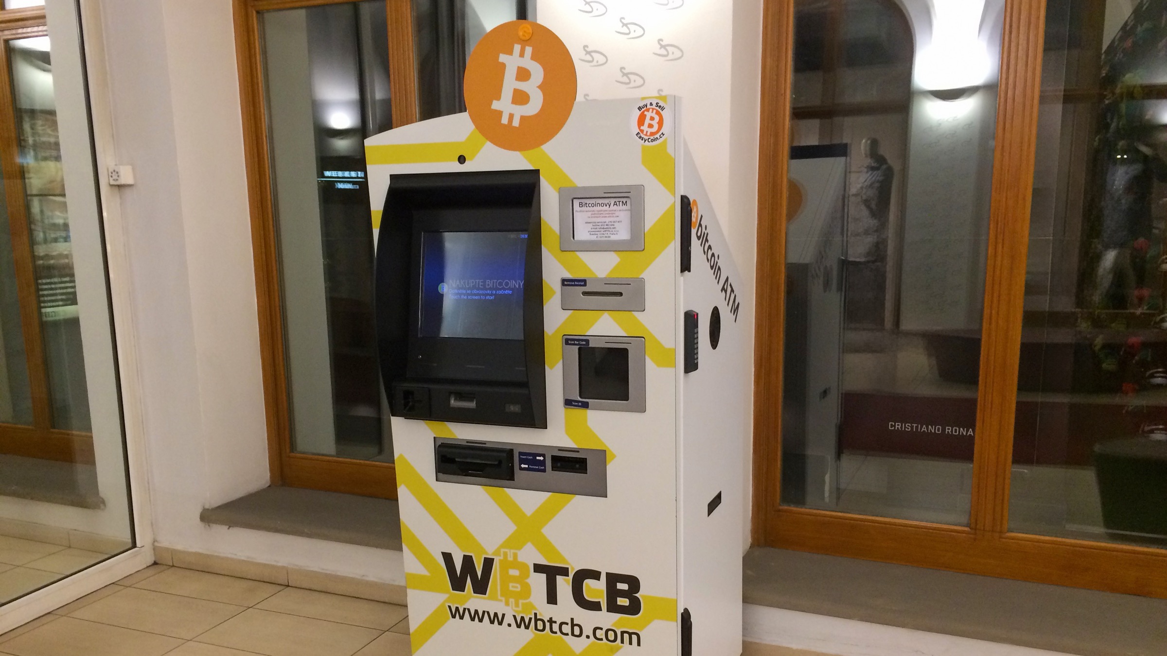 A bitcoin ATM with an orange bitcoin logo on top standing in an arcade in Prague, in front of shop windows