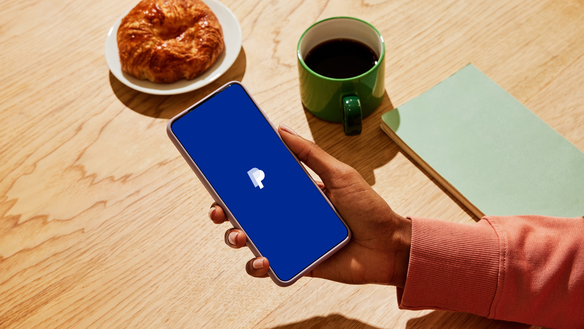 A hand holding a smartphone showing the PayPal app open on a wooden table beside a coffee cup and pastry, illustrating the app US travellers now use to scan Weixin Pay QR codes in China.