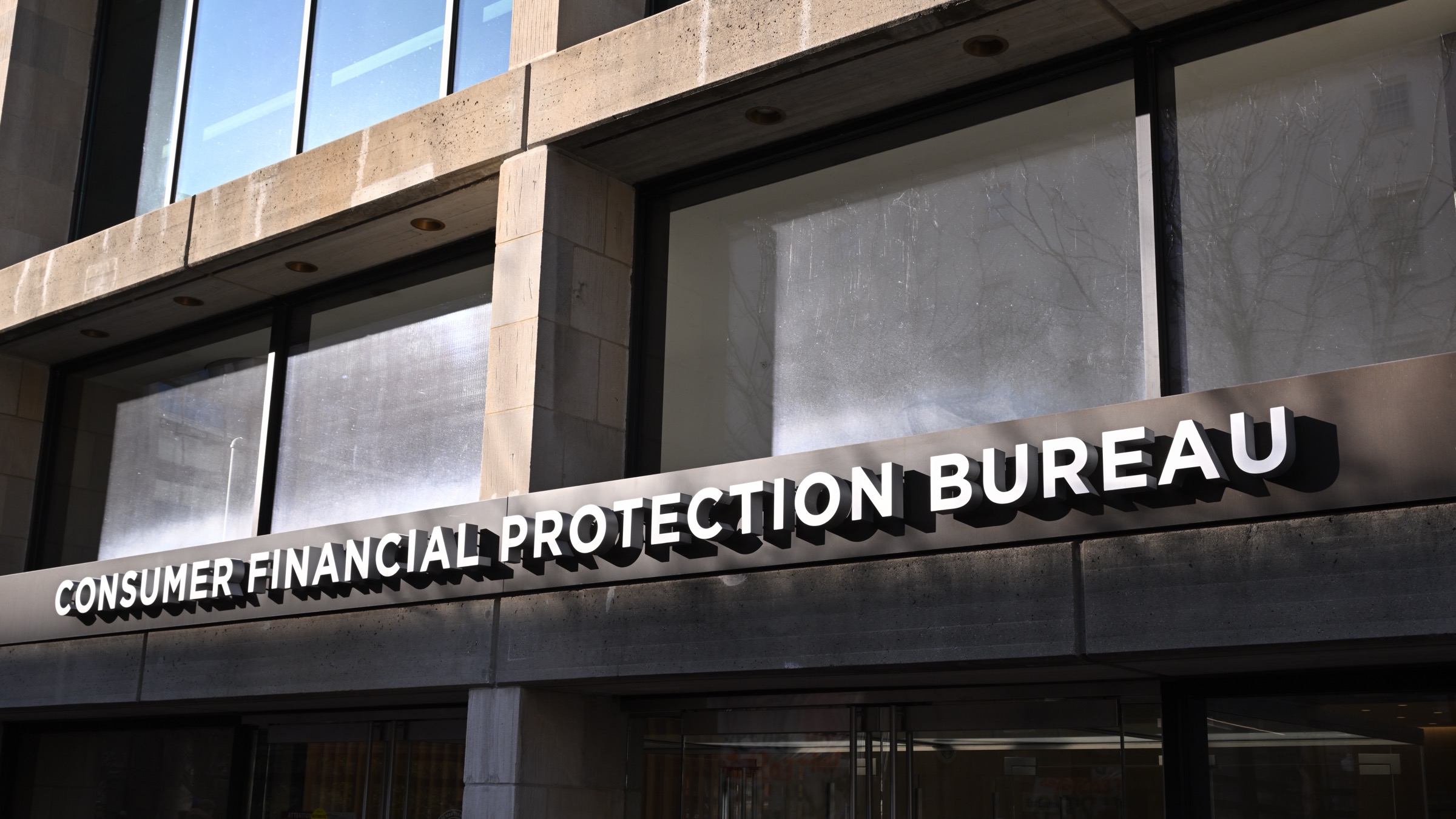 The entrance sign of the Consumer Financial Protection Bureau headquarters in Washington, with the agency name in white lettering on a dark canopy above glass doors.