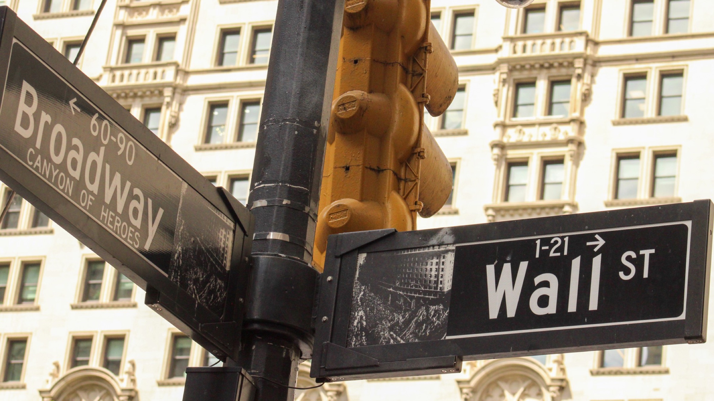 Street signs for Broadway and Wall St on a lamp post in lower Manhattan, with a traffic signal and a stone building behind them.