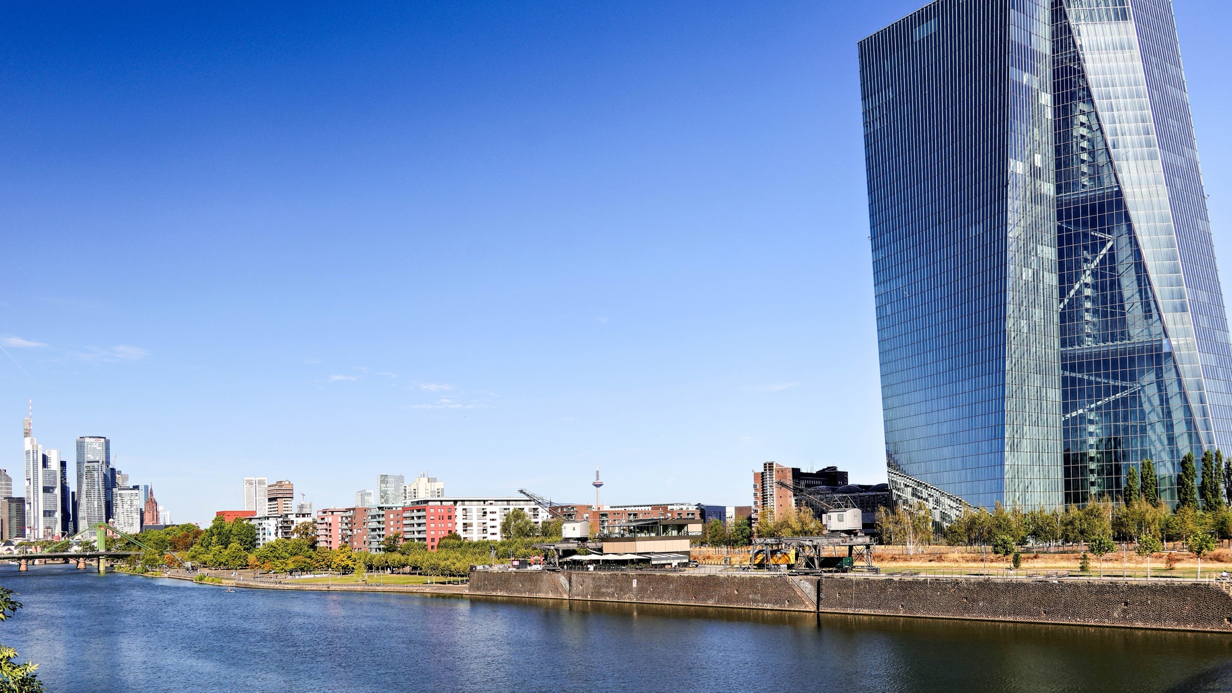 The glass twin towers of the European Central Bank beside the River Main in Frankfurt, with the city skyline in the distance under a clear blue sky.
