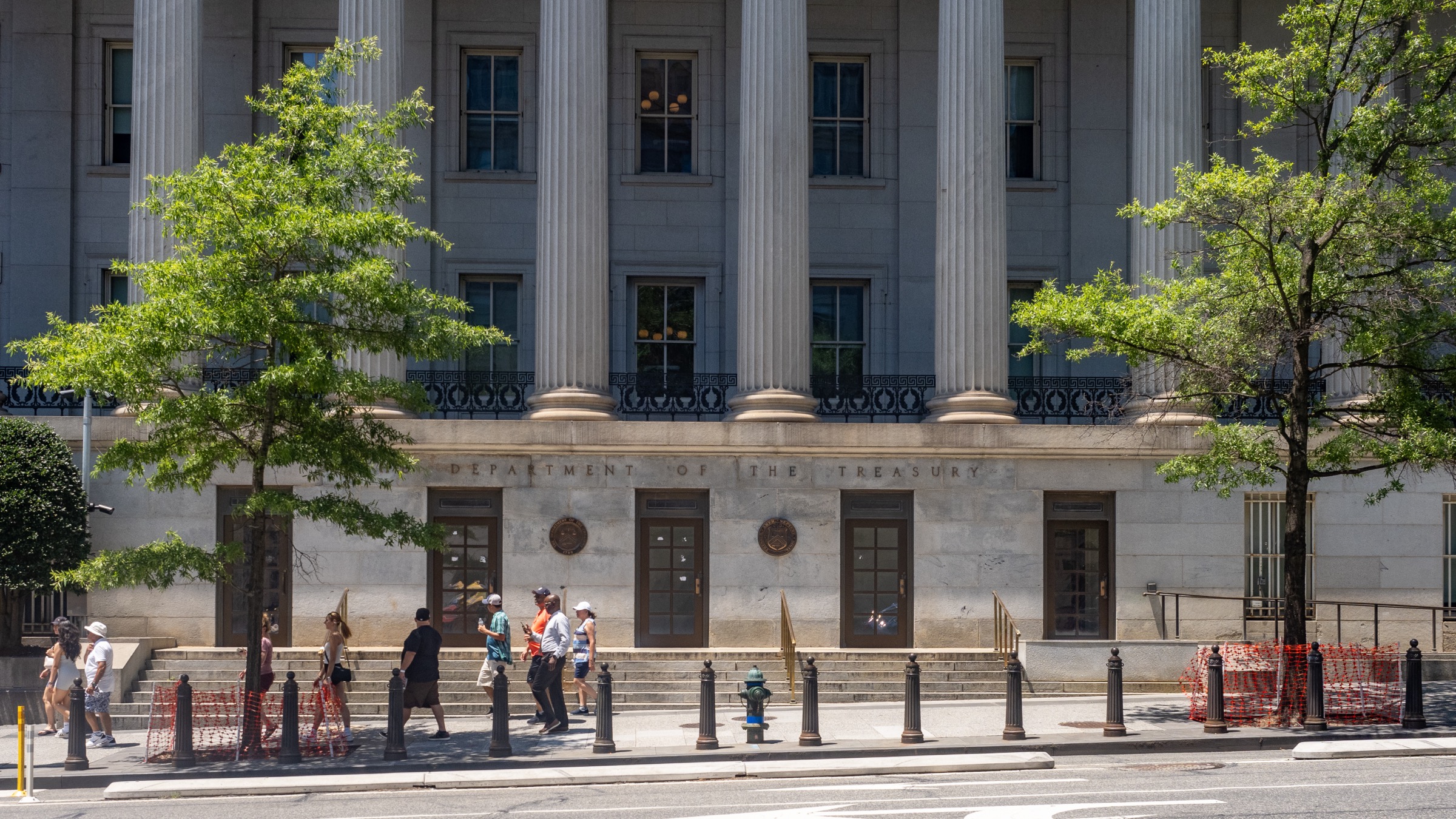 The columned south facade of the United States Department of the Treasury building in Washington, with pedestrians passing on the pavement below.