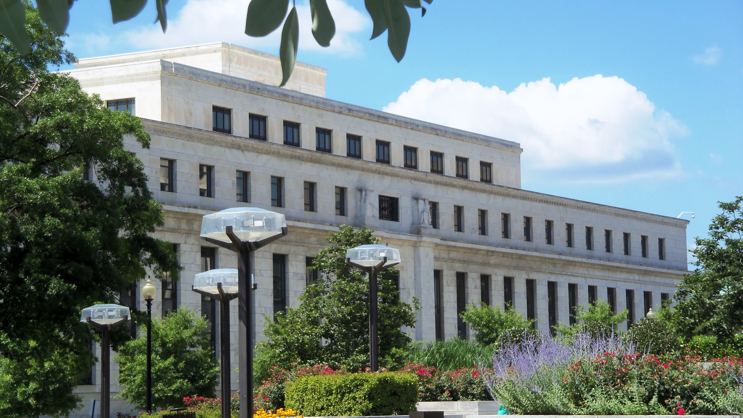 The white stone facade of the Marriner S. Eccles Federal Reserve Board Building in Washington, seen past trees and street lamps on a sunny day.