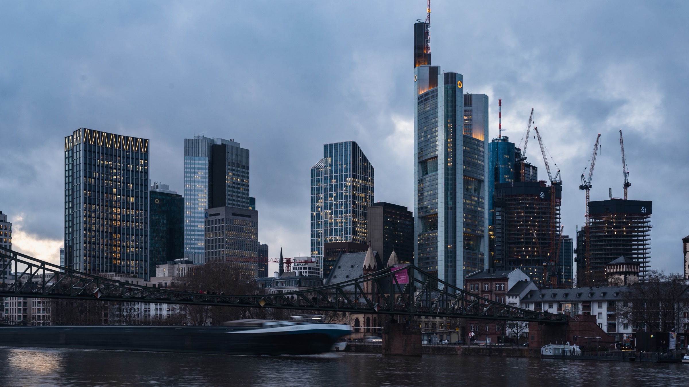 The Frankfurt banking district seen across the river Main at dusk, with the Commerzbank tower and the Eiserner Steg footbridge in the foreground.