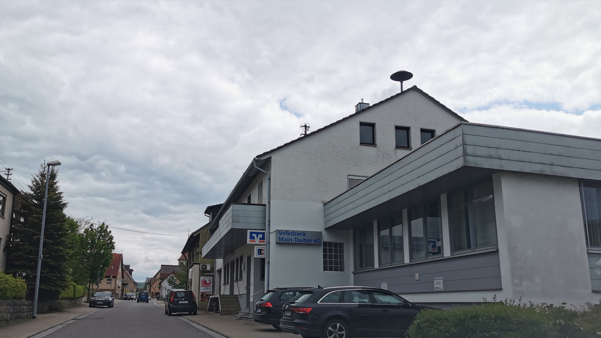 A small Volksbank branch on a village street in Germany, with the cooperative banking logo on a sign beside the entrance.