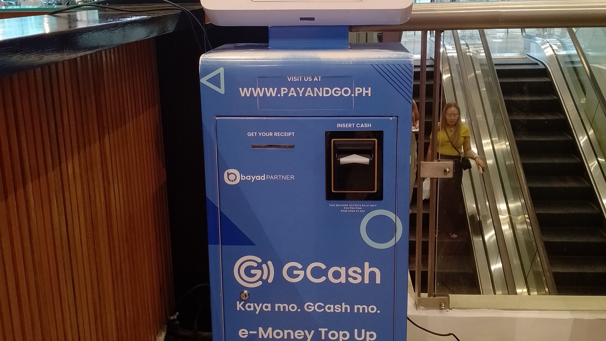 A blue GCash self-service payment kiosk beside an escalator in a shopping mall in Cebu, Philippines, marked for e-money top-up and bills payment.