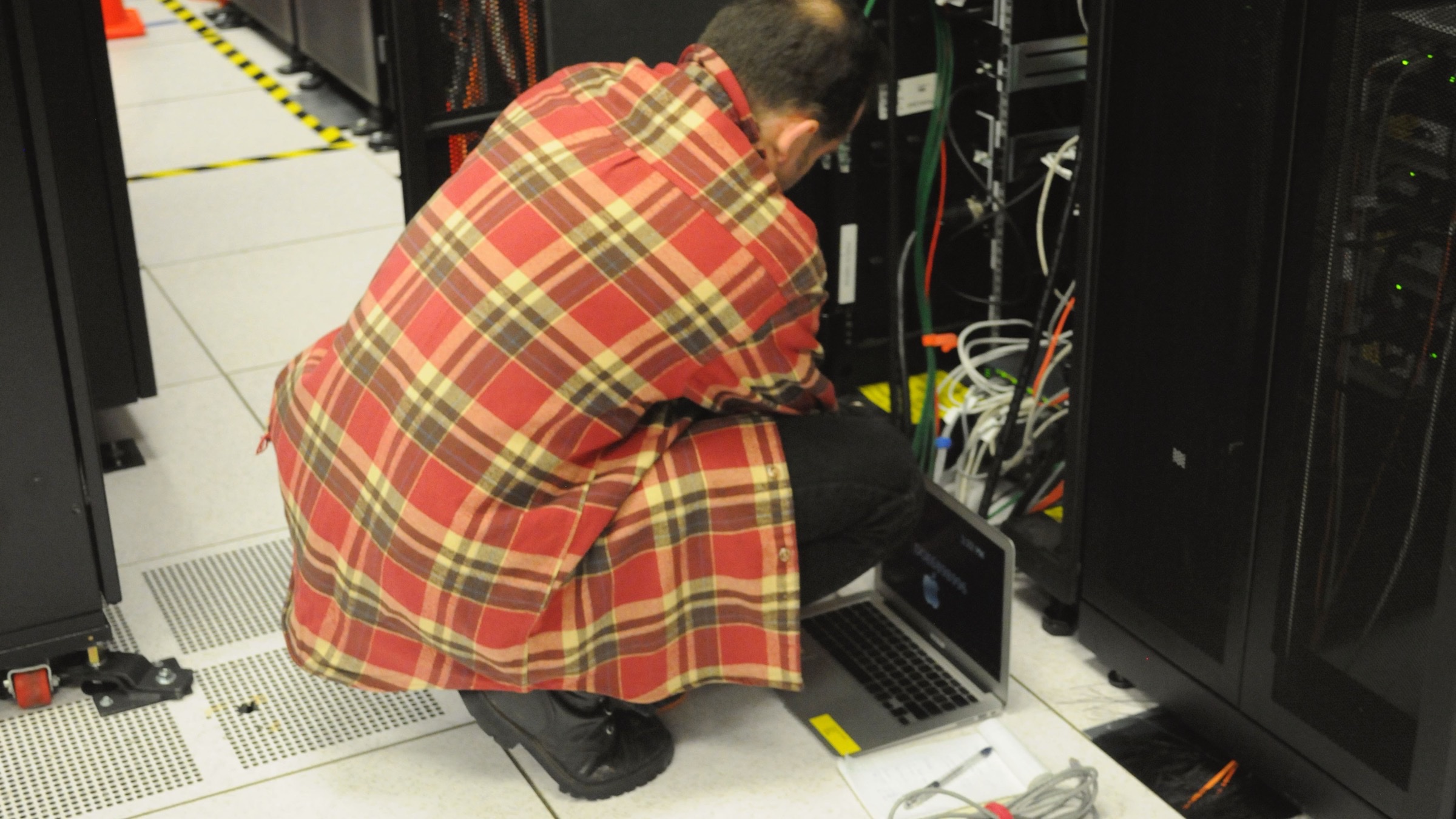 A technician crouching beside an open server rack in a data centre, working on a laptop balanced on the floor.