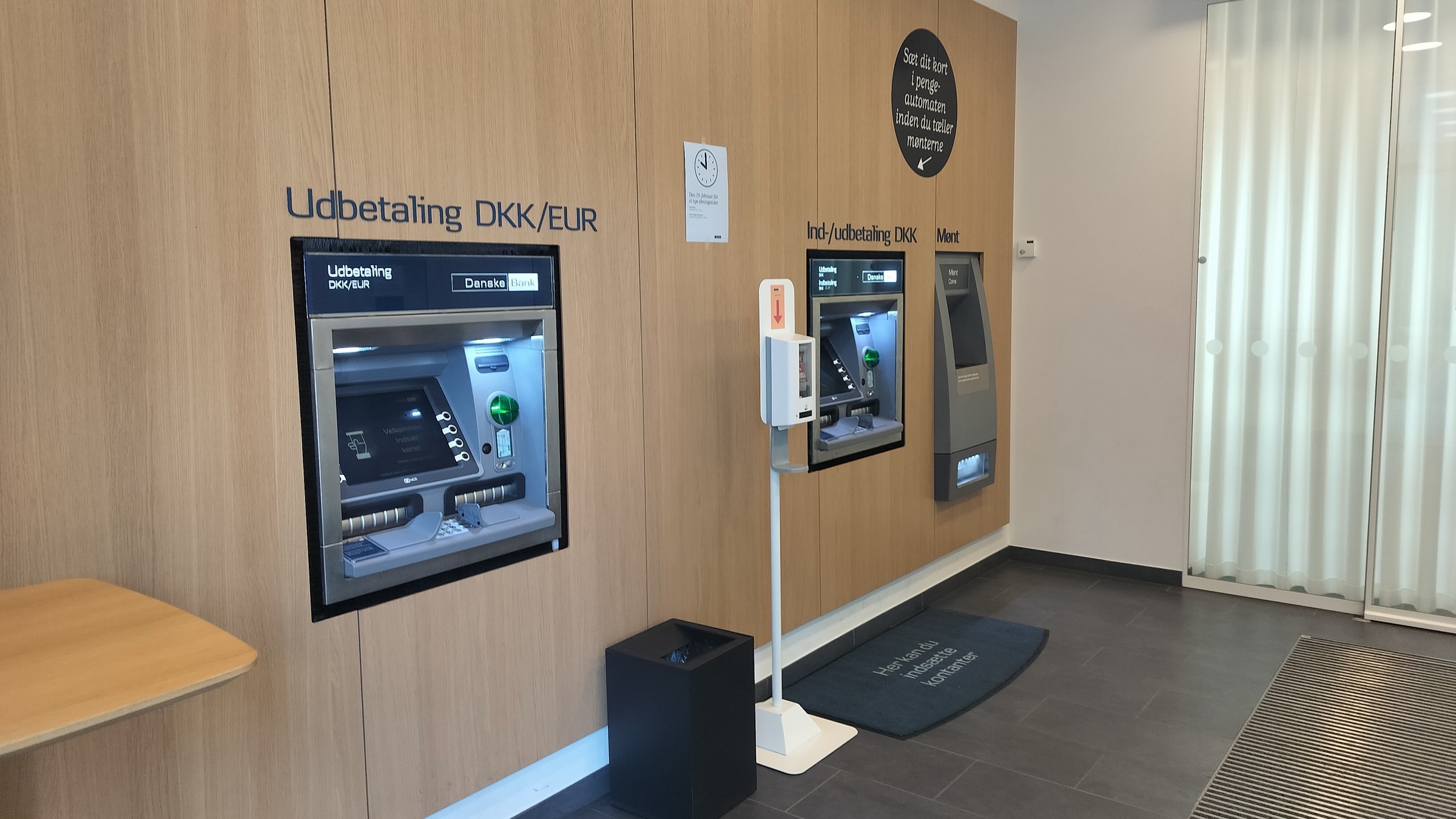 ATMs set into a wood-panelled wall in a bank lobby, with signs for cash withdrawal in Danish kroner and euros